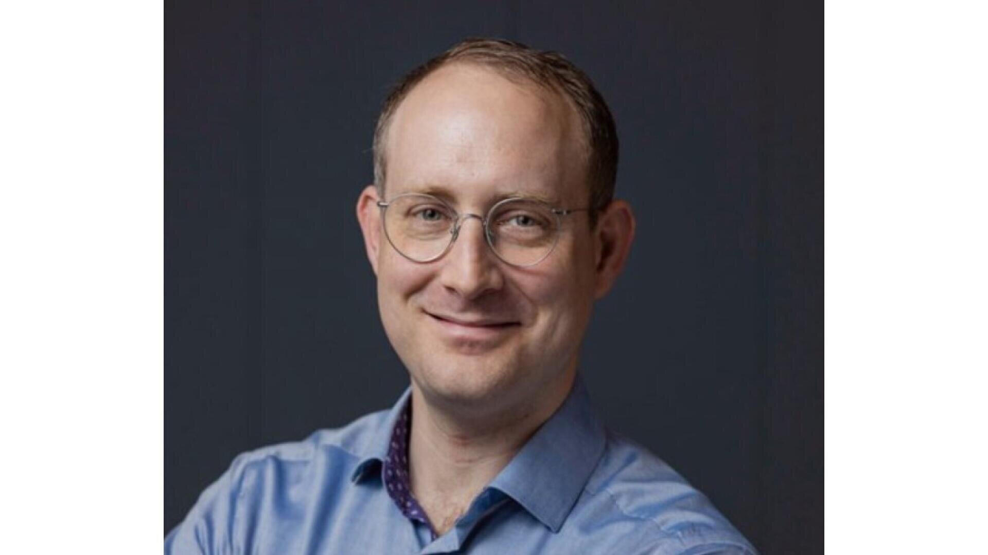 A man with short light brown hair and glasses, wearing a light blue collared shirt, smiles at the camera against a plain dark background.