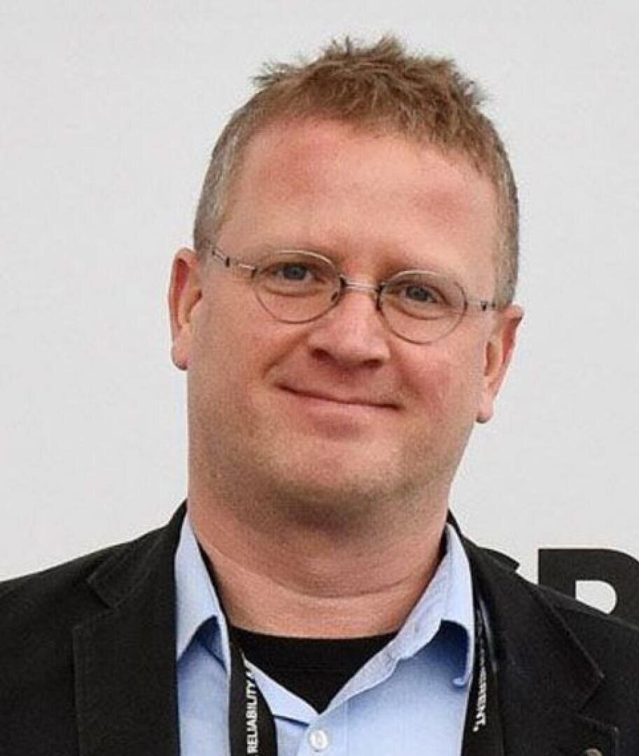 Ein Mann mit kurzen blonden Haaren und Brille, der ein hellblaues Hemd und eine schwarze Jacke trägt, lächelt vor einem hellen Hintergrund in die Kamera.