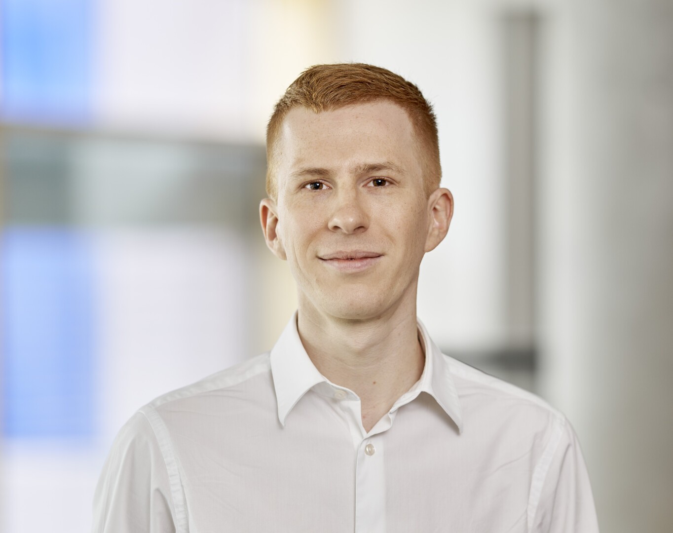 Eine junge Person mit kurzen roten Haaren, heller Haut und einem leichten Lächeln, die ein weißes Hemd mit Knöpfen trägt, steht vor einem leicht verschwommenen Innenhintergrund.