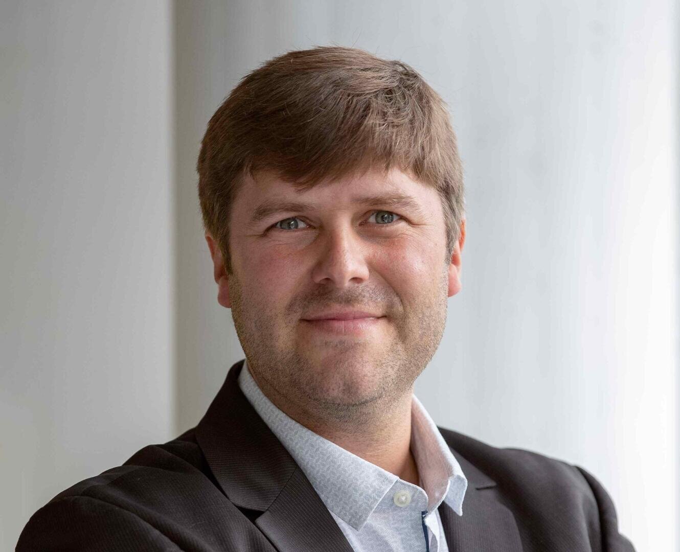 Ein Mann mit hellbraunem Haar und blauen Augen lächelt sanft. Er trägt einen dunklen Blazer über einem hellblauen Hemd mit Kragen. Der Hintergrund ist leicht verschwommen und lässt auf eine professionelle Inneneinrichtung schließen.