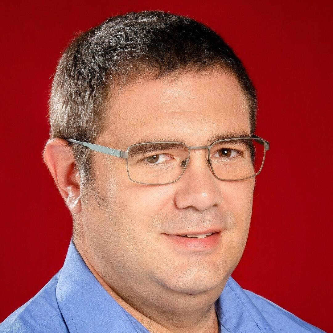 Ein Mann mit kurzen braunen Haaren, einer rechteckigen Brille und einem blauen Hemd mit Kragen lächelt leicht vor einem einfarbig roten Hintergrund.