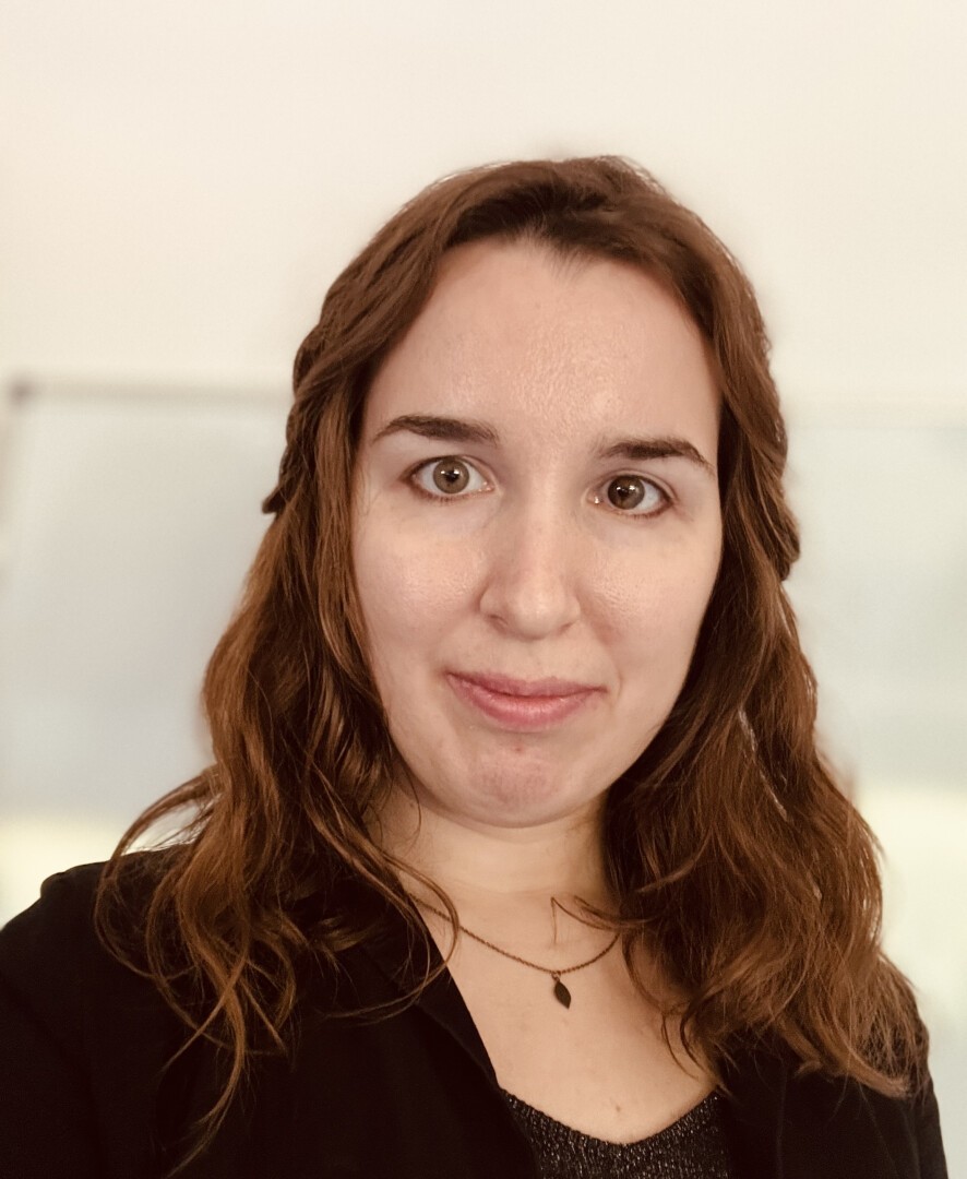 Eine Frau mit welligem braunem Haar, einem schwarzen Top und einer zarten Halskette, blickt mit einem leichten Lächeln in die Kamera. Der Hintergrund ist sanft unscharf und farblich neutral.