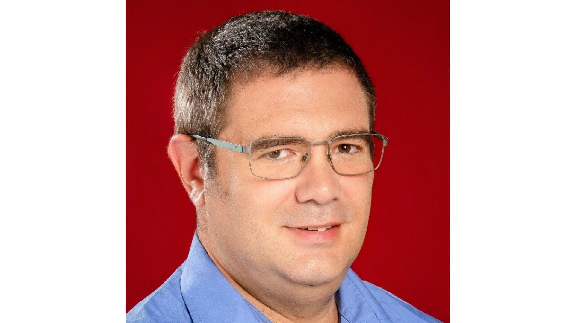 A man with short brown hair and glasses, wearing a blue collared shirt, smiles slightly in front of a solid red background.