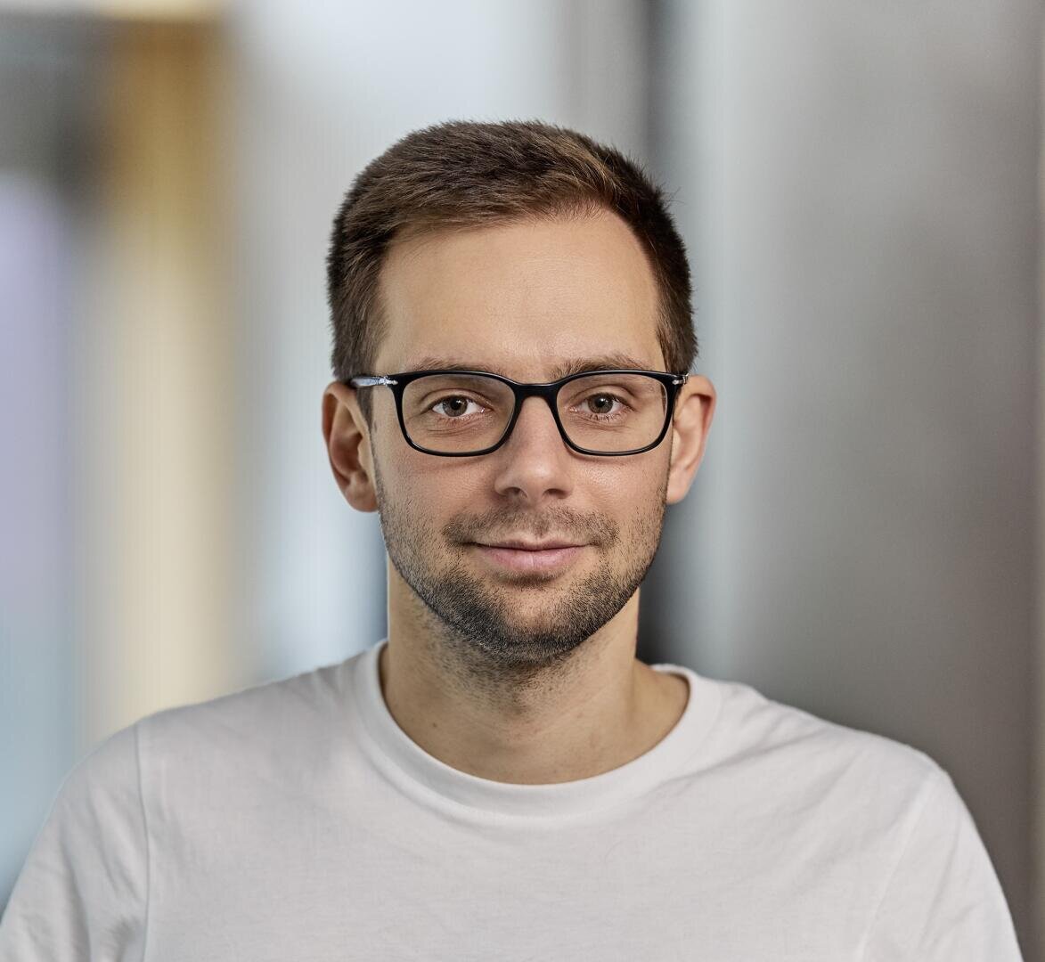 Ein Mann mit kurzen braunen Haaren, Brille und hellem Bart trägt ein schlichtes weißes T-Shirt. Er blickt in die Kamera und lächelt leicht. Der Hintergrund ist unscharf.