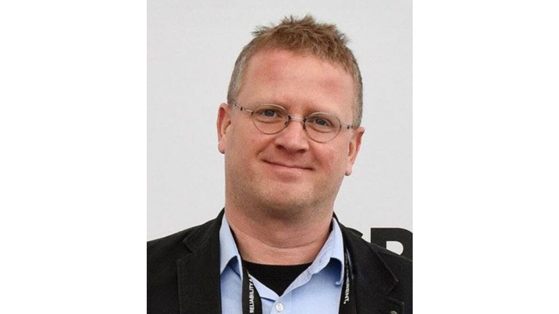 A man with short, light brown hair and glasses is smiling. He is wearing a light blue collared shirt, a black jacket, and a lanyard around his neck. The background is plain and light-colored.