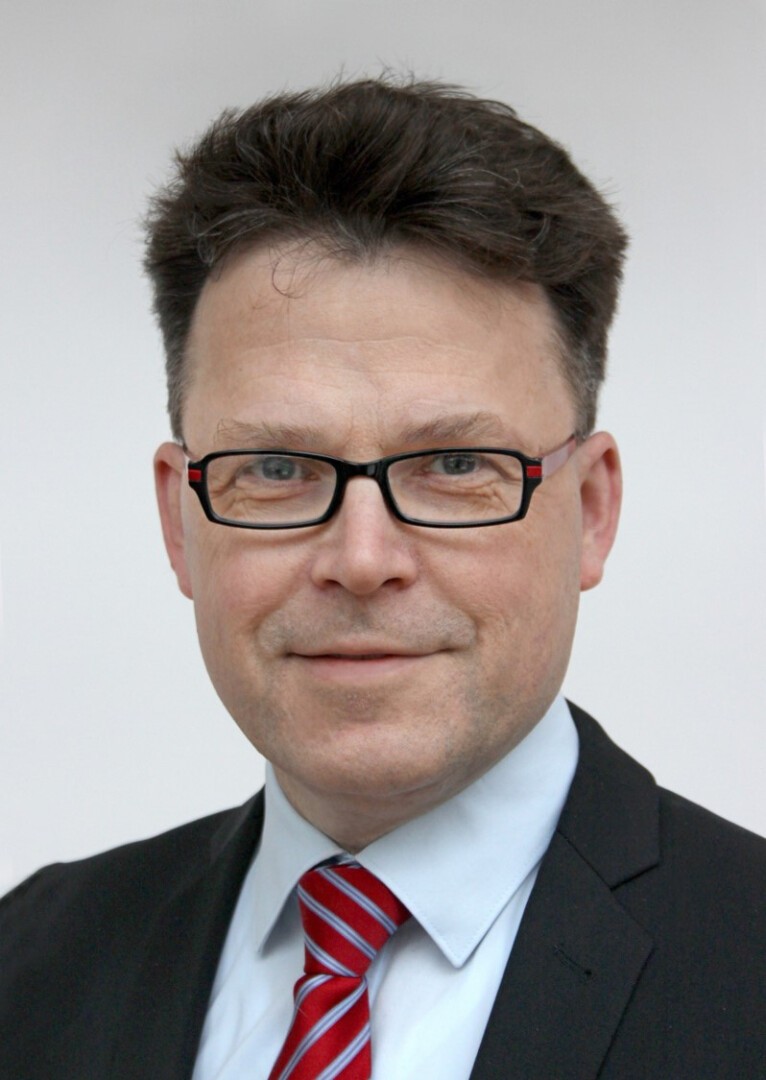 Ein Mann mit Brille, dunklem Anzug, hellblauem Hemd und rot gestreifter Krawatte, leicht lächelnd, mit kurzen dunklen Haaren und neutralem Hintergrund.