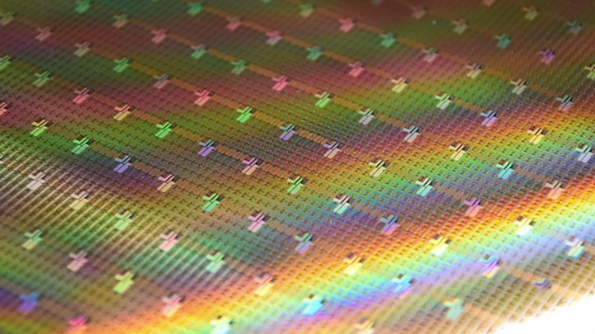 Nahaufnahme eines Siliziumwafers mit Mikrochips, der ein Gitter aus winzigen regenbogenfarbenen Quadraten zeigt, die das Licht in verschiedenen Farben reflektieren und so ein leuchtendes und schillerndes Muster erzeugen.