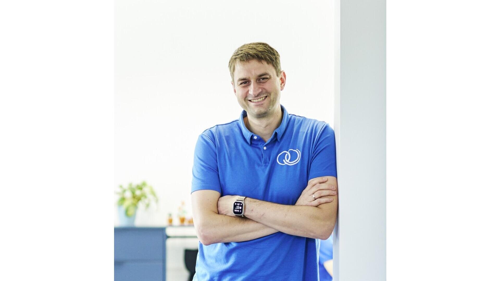 A man wearing a blue polo shirt with a white logo stands indoors, leaning against a white wall with arms crossed and smiling. A plant and blue furniture are blurred in the background.