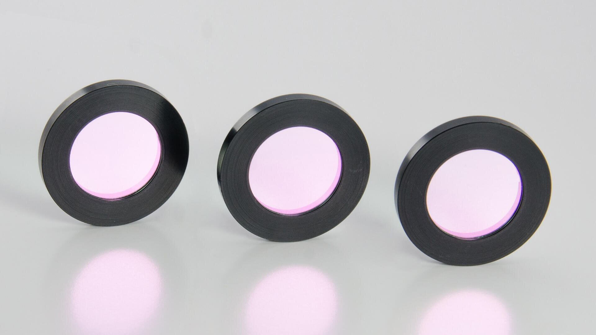 Three circular optical filters with black metal rims and faintly purple-tinted glass centers are standing upright on a white surface, casting soft reflections below them.