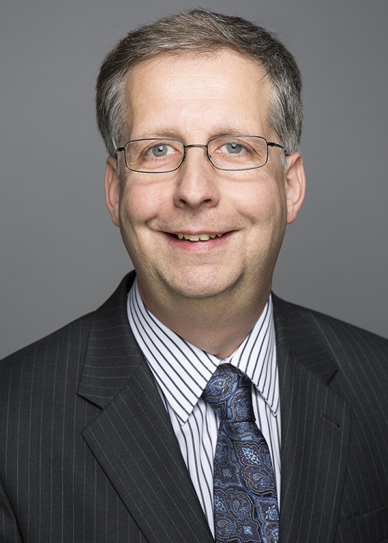 Ein Mann mittleren Alters mit kurzen grauen Haaren, Brille und einem gestreiften Jackett über einem weißen Hemd mit dünnen Streifen und einer gemusterten blauen Krawatte, der vor einem schlichten grauen Hintergrund lächelt.