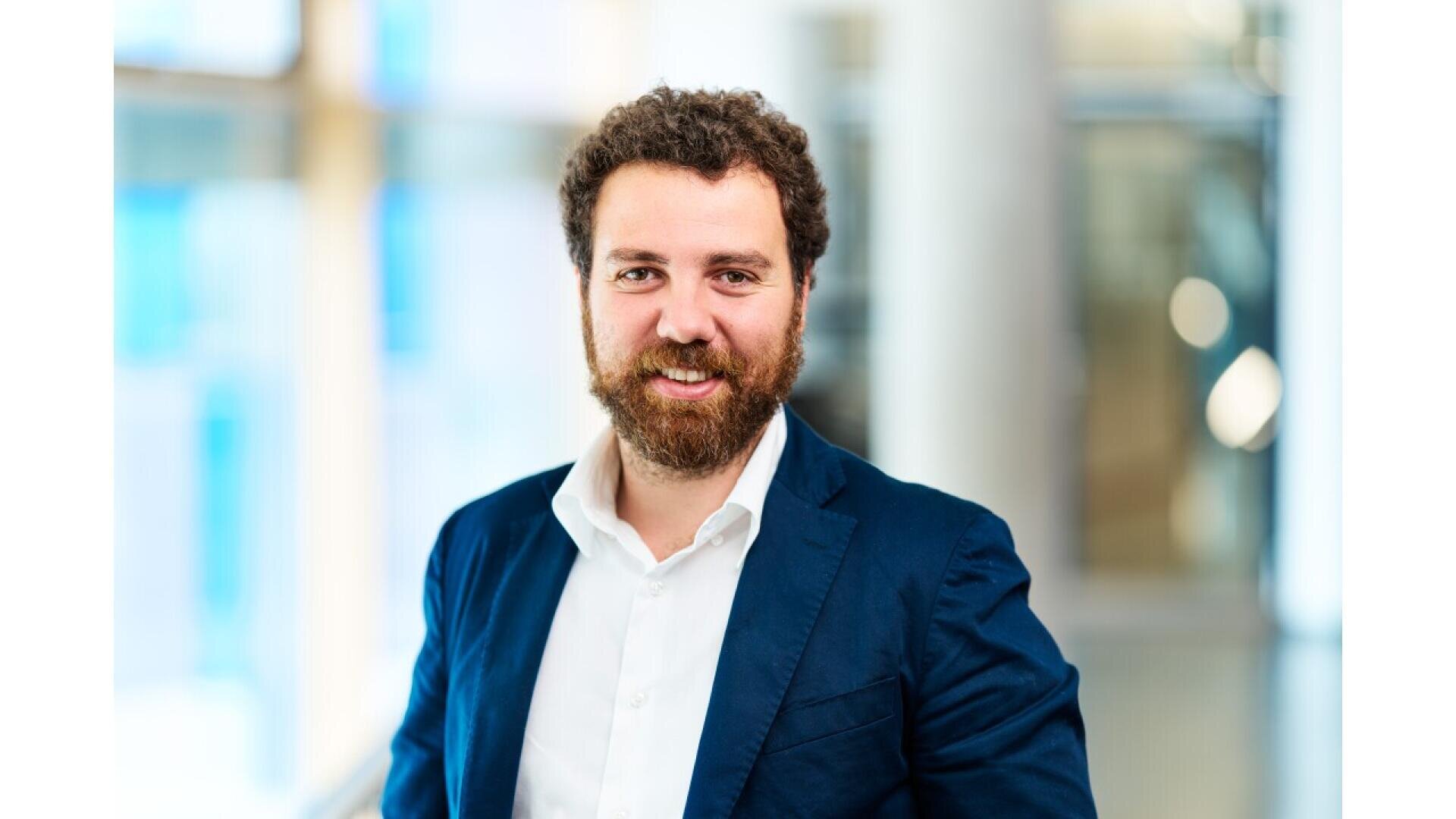 A man with curly brown hair and a beard, wearing a navy blue blazer and white shirt, smiles while standing indoors in a brightly lit, modern setting.