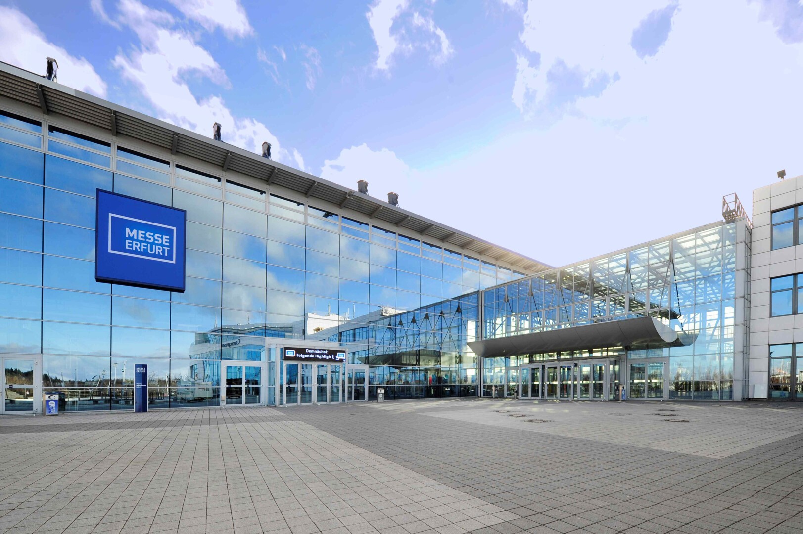 The image shows the exterior of Messe Erfurt, a modern glass-fronted convention and exhibition center with a large blue sign and wide paved entrance under a partly cloudy sky.