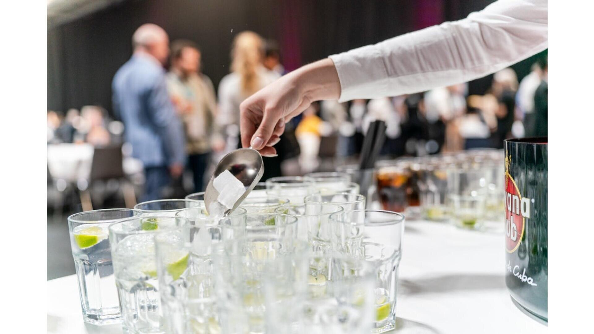 A person uses tongs to add ice cubes to a row of clear glasses filled with drinks and lime slices at an event, with blurred people in the background.