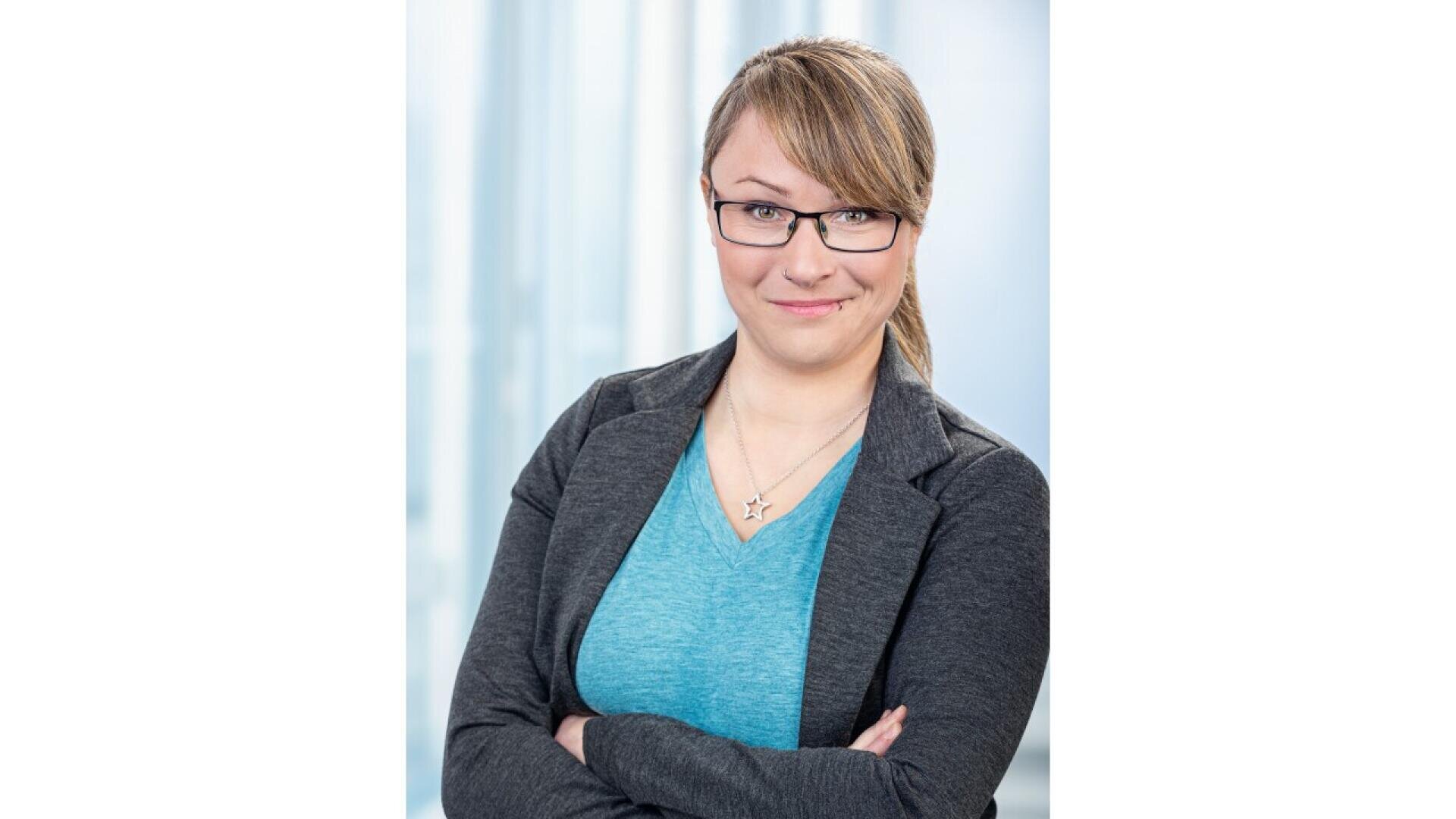 A woman with light brown hair, glasses, and a slight smile stands with arms crossed, wearing a gray blazer over a blue top, in front of a blurred light background.