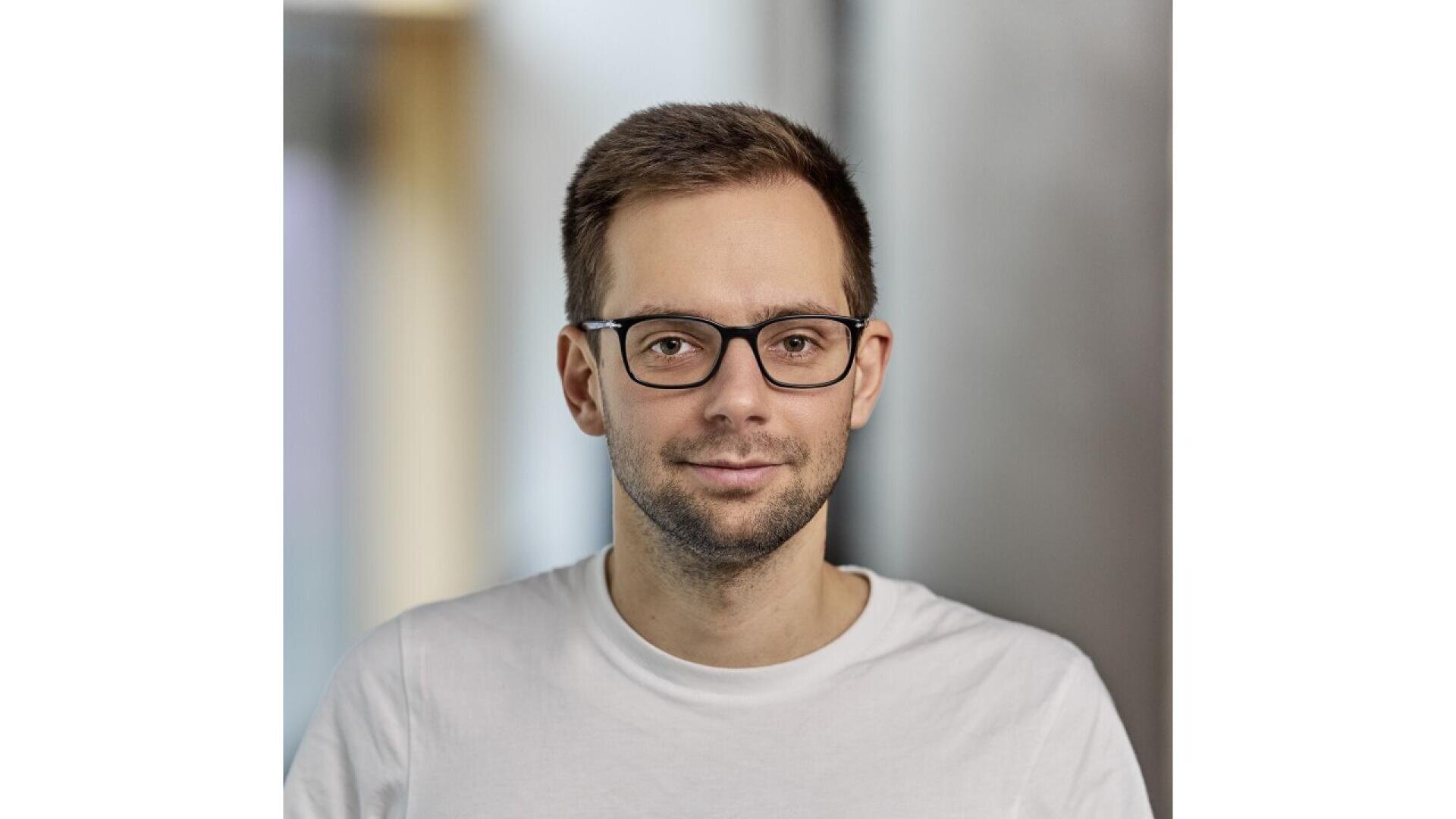 A man with short brown hair and glasses, wearing a white T-shirt, is looking at the camera with a neutral expression. The background is softly blurred.