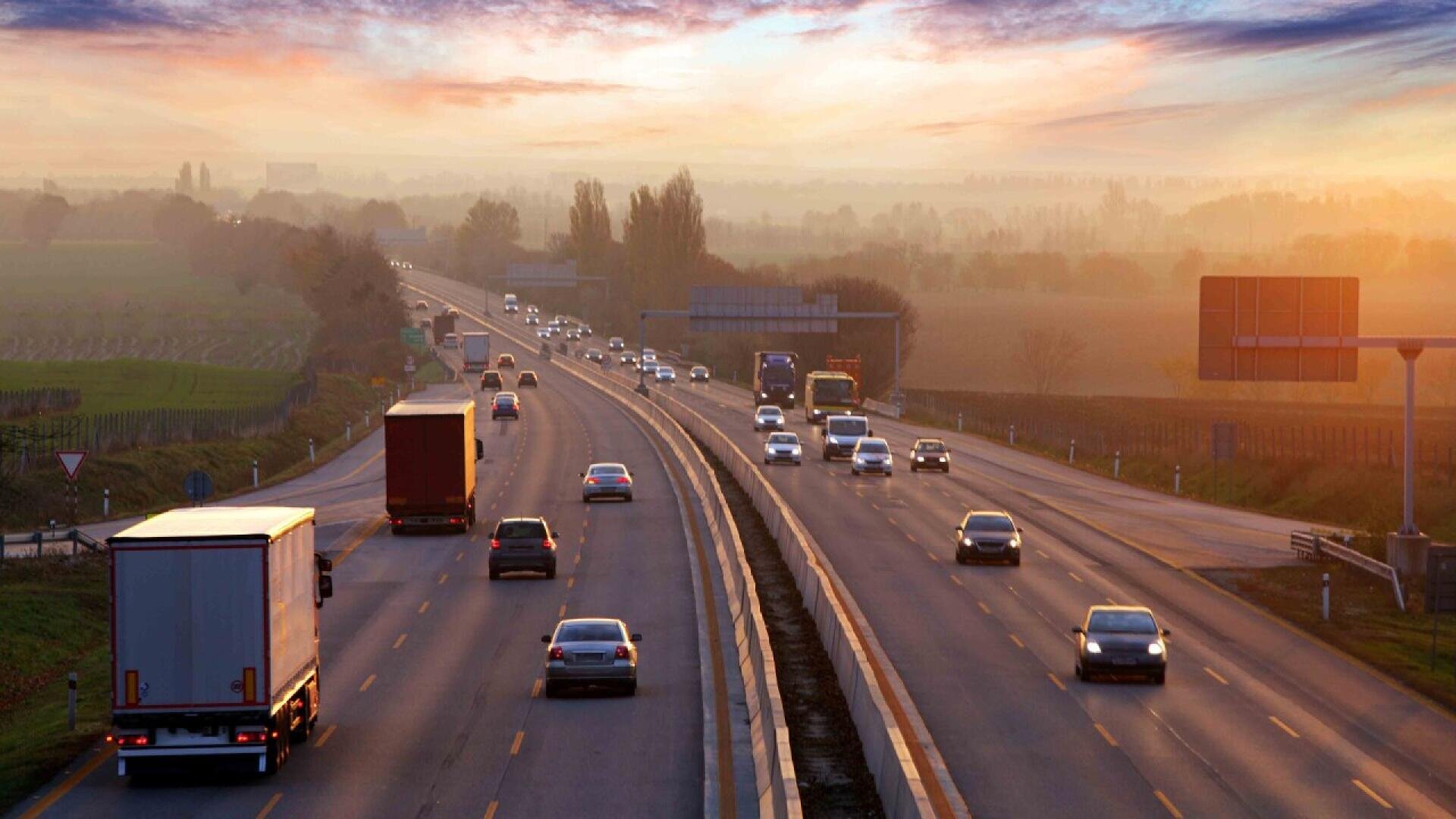 Cars and trucks drive on a multi-lane highway at sunset, with fields and trees on both sides and a colorful sky in the background.