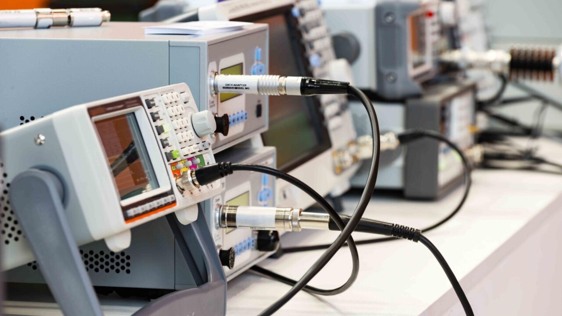 A row of electronic testing equipment, including oscilloscopes and signal generators, connected with various cables on a laboratory bench.