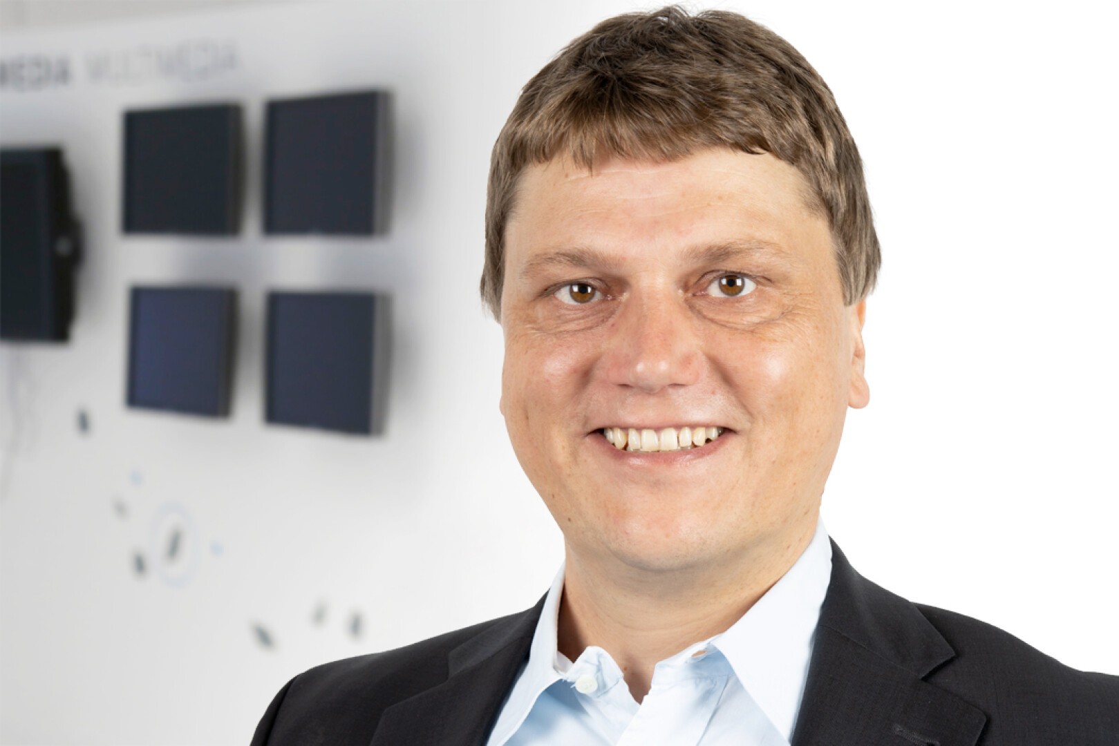 Ein lächelnder Mann im Jackett und hellblauem Hemd steht vor einer unscharfen Wand mit kleinen schwarzen Bildschirmen und schwachem Text im Hintergrund.