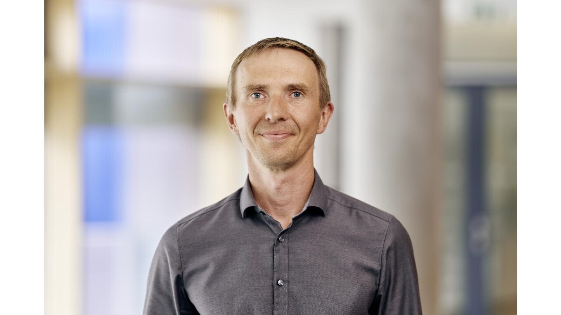 A man with short light brown hair, wearing a grey button-up shirt, stands indoors in front of a blurred background with neutral colors. He is facing the camera and smiling softly.