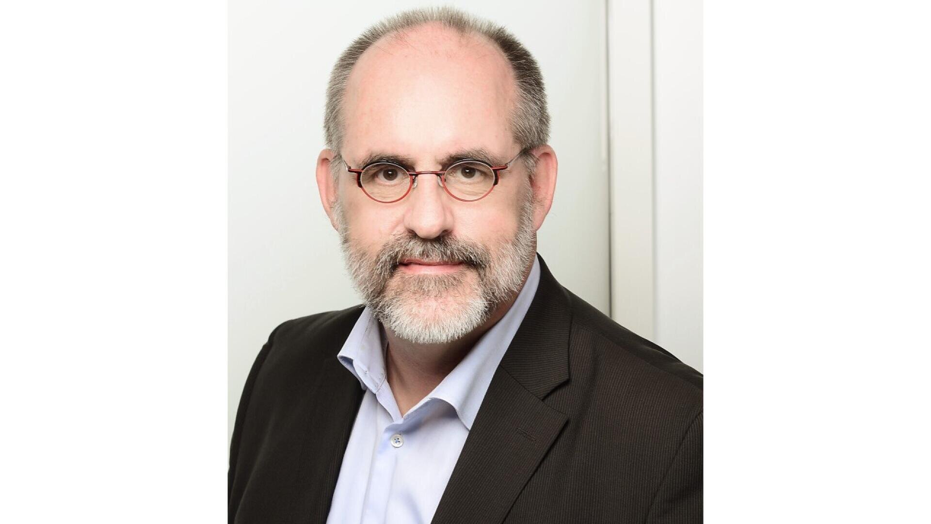 A middle-aged man with a beard and glasses, wearing a dark suit jacket over a light blue collared shirt, poses in front of a plain white background.