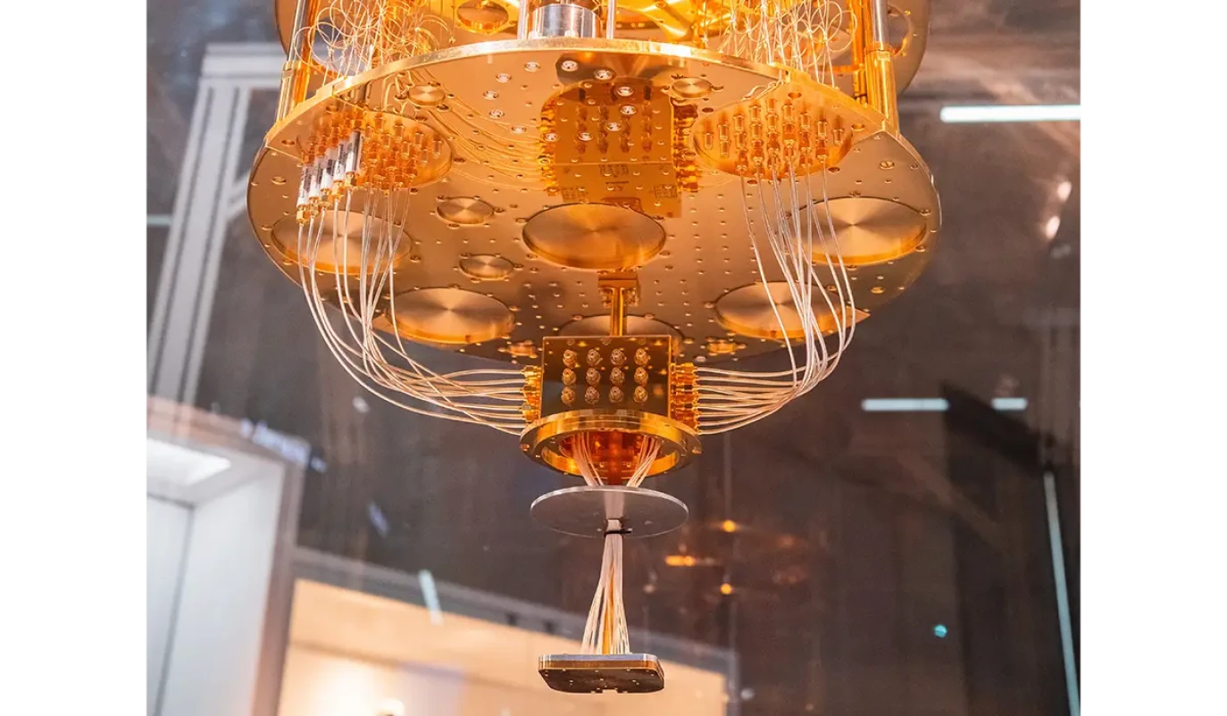 Eine goldfarbene, kreisförmige Kühlstruktur eines Quantencomputers hängt von oben herab. Zahlreiche dünne Kabel verlaufen von der Hauptstruktur zu einem kleineren Bauteil am unteren Ende. Im Hintergrund sind Glasflächen und eine Laborumgebung erkennbar.