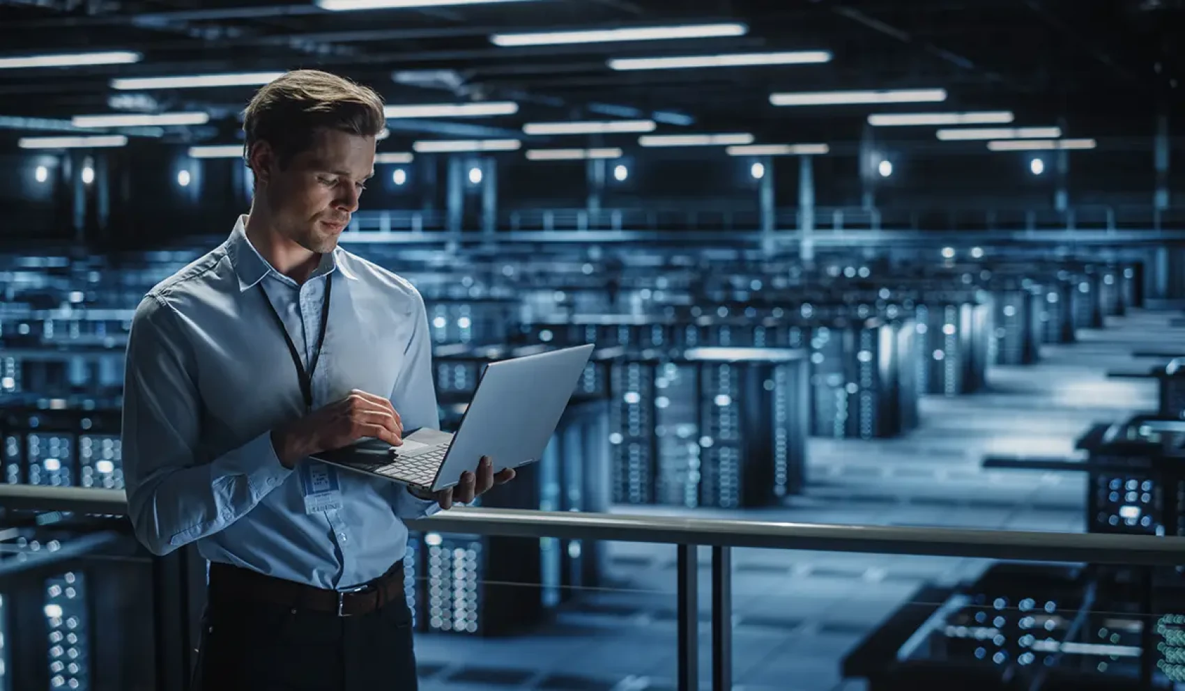 A man with a laptop stands on a gallery above a large room full of server racks and modern computer technology.